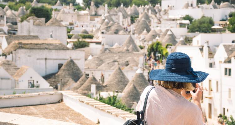 Een vrouwelijke reiziger in een brede blauwe hoed fotografeert de kegelvormige trulli-daken van Alberobello.