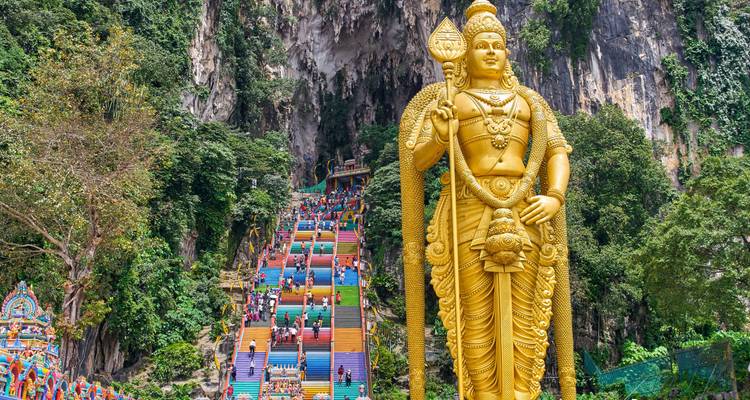 La statue dorée du Seigneur Murugan se dresse devant l'escalier coloré menant aux grottes de Batu, avec des visiteurs qui y montent.