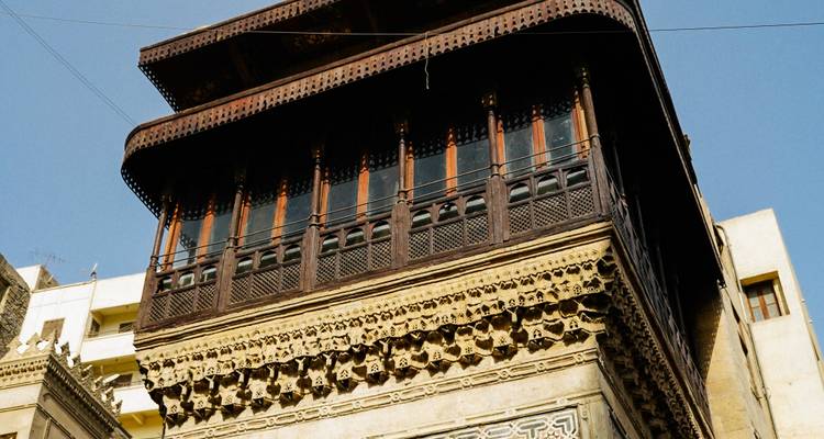 Upward view of an intricately carved wooden mashrabiya balcony on a historic building against a clear sky.