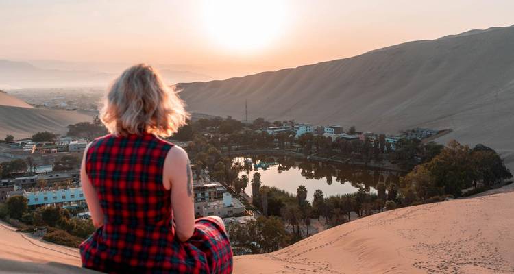 Traveler in a red plaid dress sits atop a sand dune overlooking the Huacachina desert oasis at sunset.