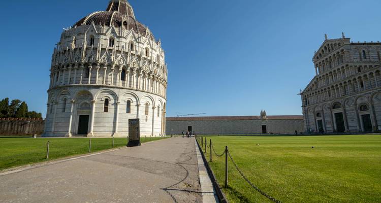 Wide view of Pisa’s Baptistery and Cathedral across a green lawn beneath a clear blue sky.