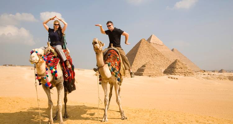 Dos turistas posando de manera divertida en camellos frente a las pirámides bajo cielos nublados