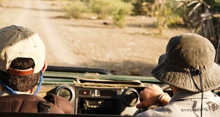 Two safari guides drive an open jeep along a dusty track in the African bush.