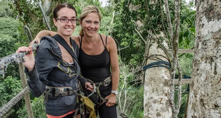 Dos mujeres sonrientes con arneses de seguridad de pie en una plataforma de árbol muy por encima de la jungla.