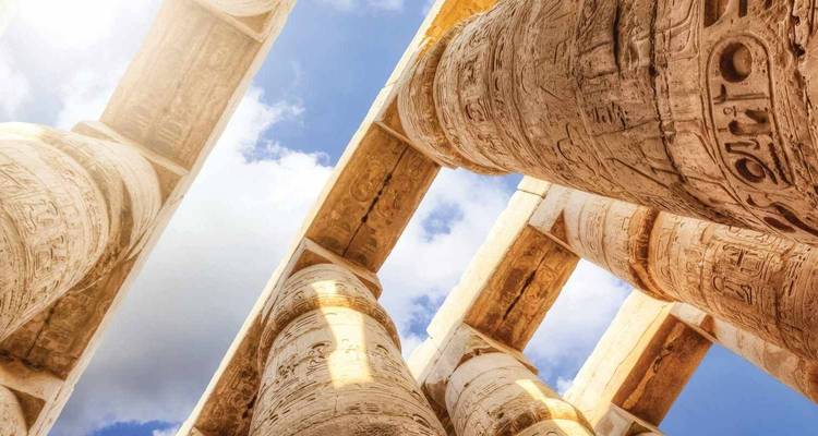 D'immenses colonnes de pierre ornées de hiéroglyphes gravés encadrent des portions de ciel bleu au-dessus.
