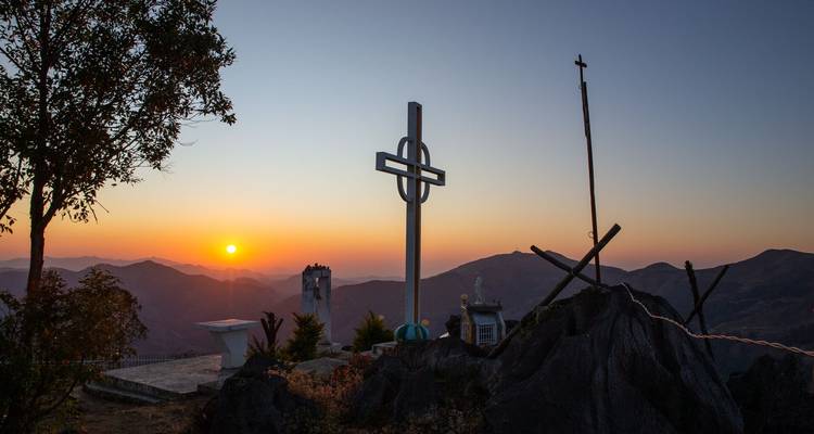 Mountain summit scene at sunrise featuring a large cross monument overlooking distant peaks.