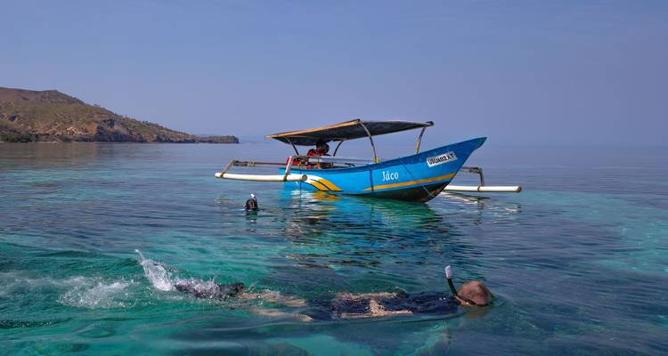 Snorkelers explore clear turquoise water near a small blue wooden boat against a calm sea backdrop.