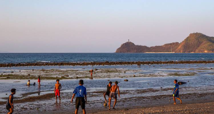 Local boys play soccer on a sandy beach at low tide with ocean and hills in the distance.