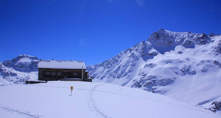 Snow-covered alpine hut standing alone against bright blue sky and high mountain peaks.