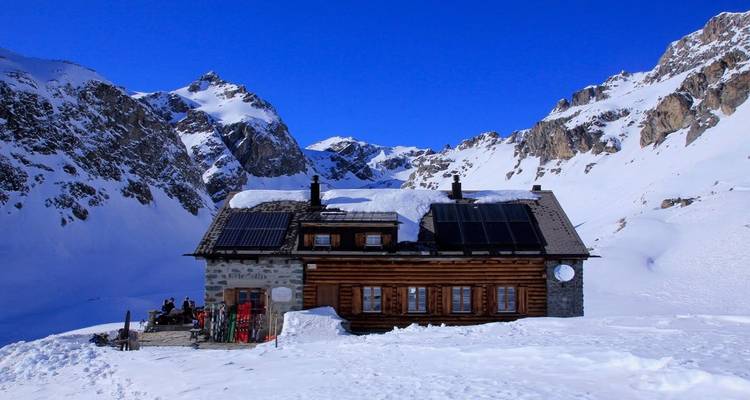 Log mountain refuge with solar panels surrounded by snowy peaks and skiers relaxing outside.