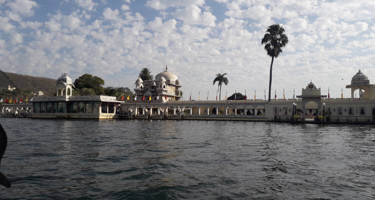 Lake palace complex and palm tree reflected in the waters of Udaipur’s Lake Pichola beneath a sky of scattered clouds.