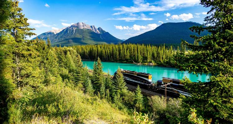 Un train de luxe pour visites touristiques serpente devant une rivière d'un turquoise éclatant au milieu de forêts de pins denses et de sommets imposants des Montagnes Rocheuses sous un ciel bleu dégagé.