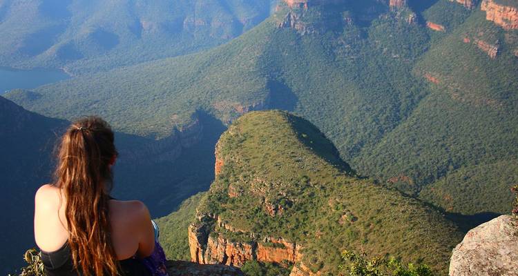 Een jonge reiziger zit op de rand van een klif en kijkt uit over de uitgestrekte groene heuvels en rivier van de Blyde River Canyon.