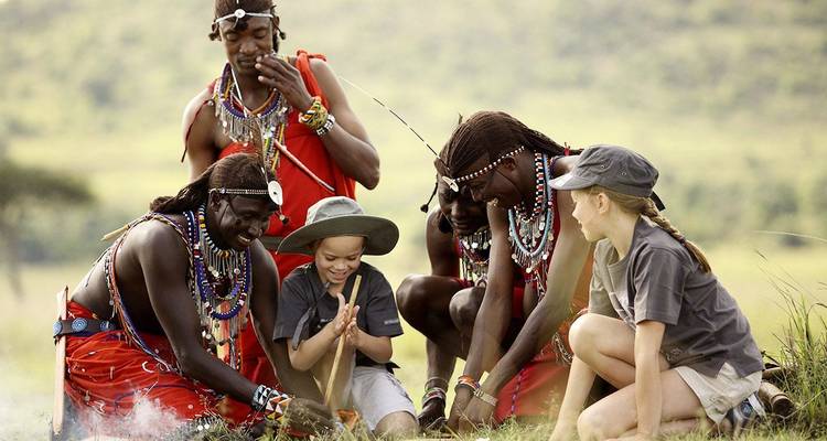 Maasai tribesmen teach two visiting children how to start a fire on open grassland.