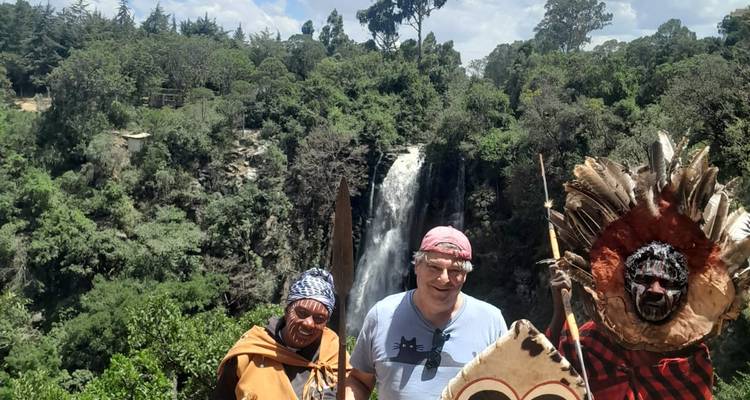 Visitors in traditional attire and tourist posing before a lush waterfall in forested hills