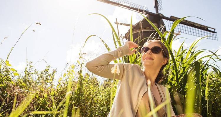 Smiling woman wearing sunglasses sits in tall grass with a traditional Dutch windmill behind her under bright sunlight.