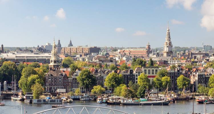 Panoramic view of Amsterdam's historic canal waterfront and church towers beneath a clear blue sky.