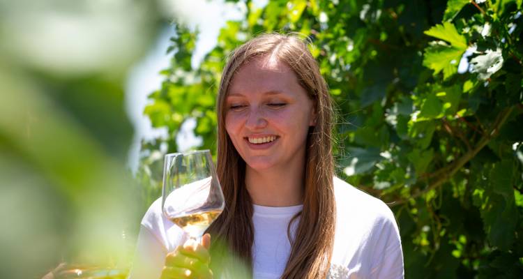 Une femme sourit en examinant un verre de vin blanc parmi le feuillage luxuriant d'un vignoble.