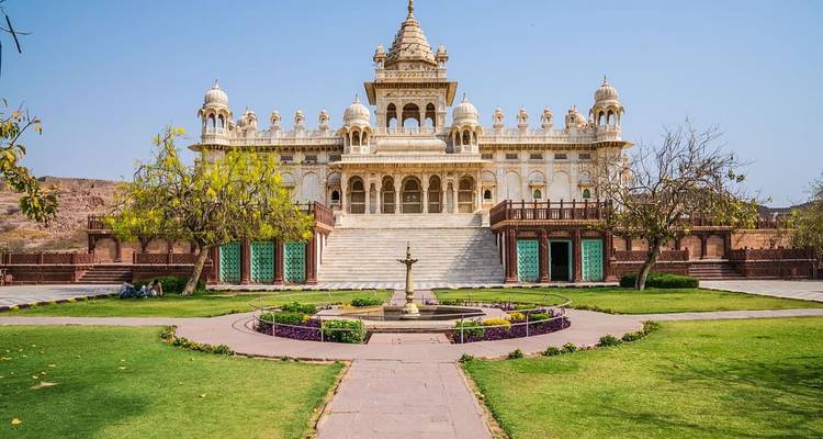Elegant white marble palace with domes and arches, fronted by a circular garden and backed by arid hills under a bright blue sky.