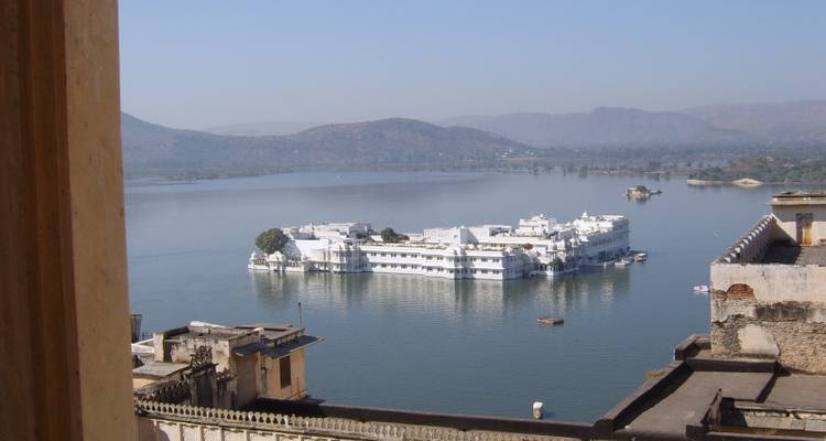 Serene lake with a white palace floating at its centre, viewed from a high stone window, with hazy hills in the background.