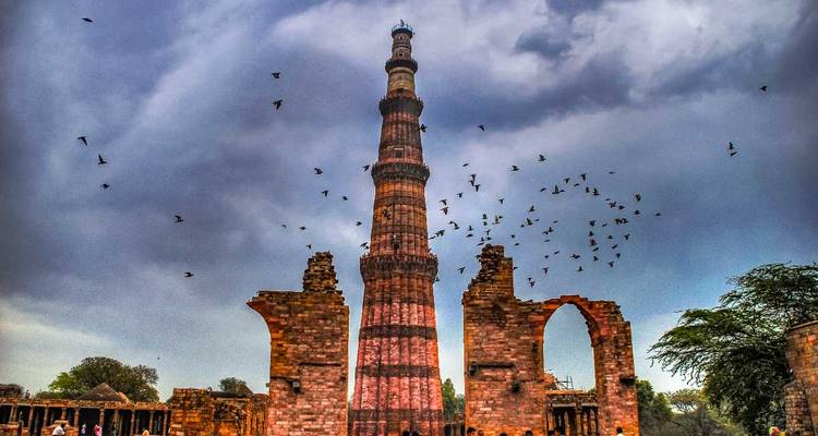 Soaring red-sandstone minaret of Qutub Minar framed by ruined arches and a flock of birds against a moody sky.