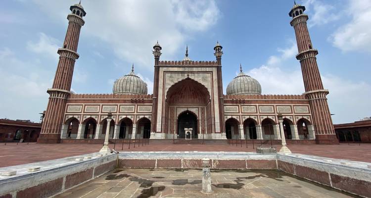 Grand façade of Jama Masjid with three domes and two tall minarets under a bright sky, reflected in a foreground ablution pool.
