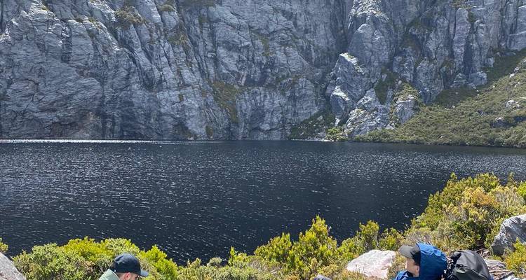 Twee wandelaars die uitrusten aan een donker alpien meer onder hoge grijze rotsen in Tasmanië.