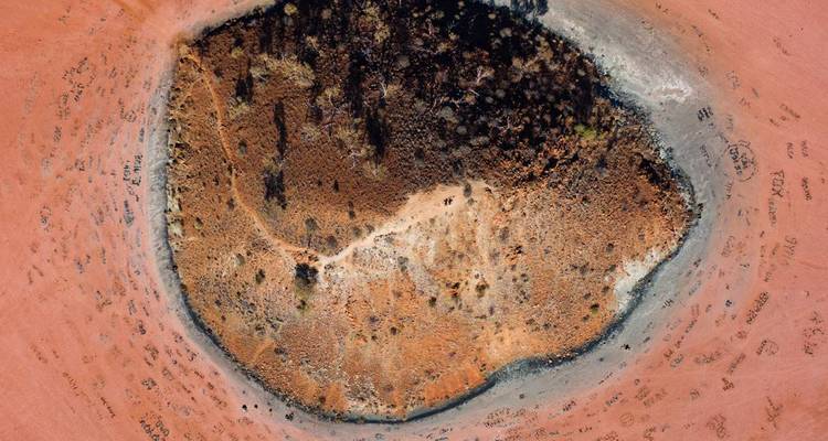 Drohnenfoto einer kreisförmigen kratähnlichen Formation, umgeben von rotem Wüstensand mit eingeritzten Besucherspuren.
