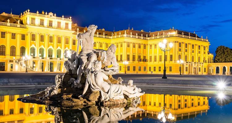 Das beleuchtete Schloss Schönbrunn mit einem Springbrunnen, der sich im Wasser spiegelt.