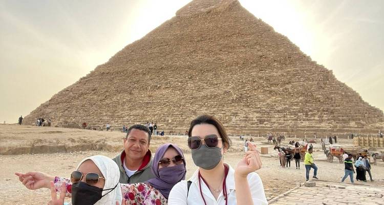Four masked travellers pose for a selfie in front of the Great Pyramid of Giza on a hazy sunny afternoon.
