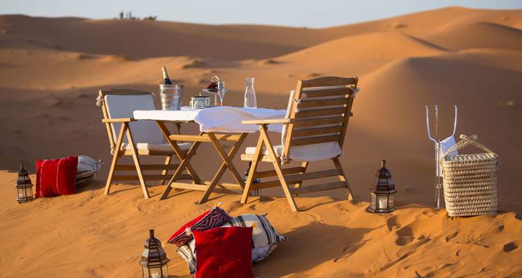 Elegante picknicktafel met kussens en lantaarns gerangschikt op gouden woestijnduinen bij zonsondergang.
