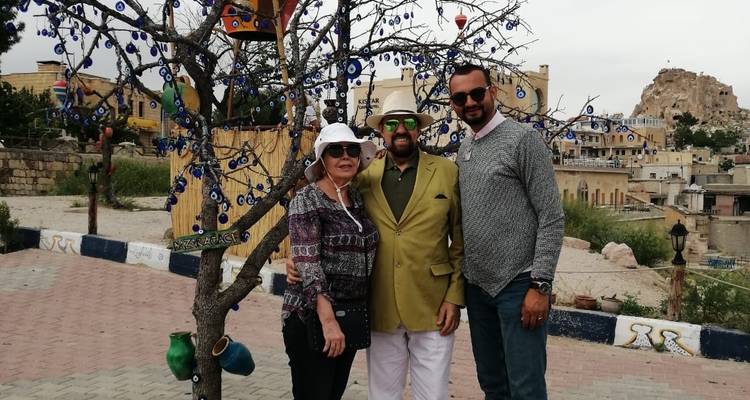 Three people standing by a tree decorated with blue talismans, and a rocky hill in the background.