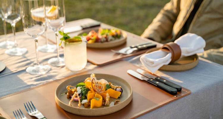Comida gourmet servida al aire libre en una mesa puesta con cubiertos y cristalería.