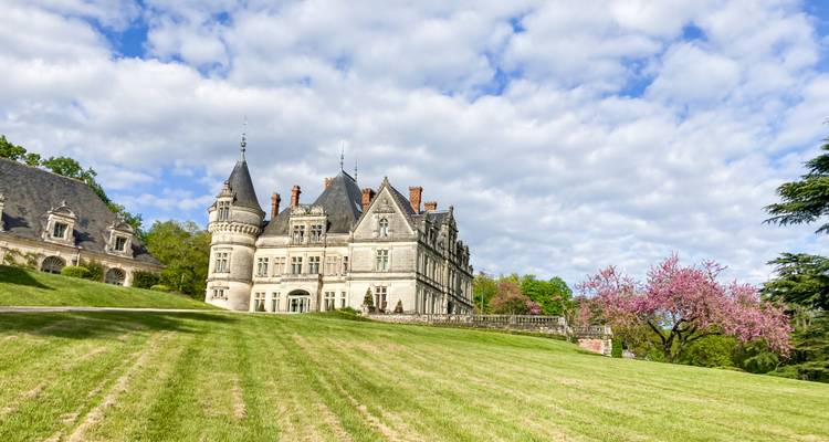 Un château majestueux aux tourelles se dresse sur une pelouse verte impeccablement entretenue sous un ciel partiellement nuageux.