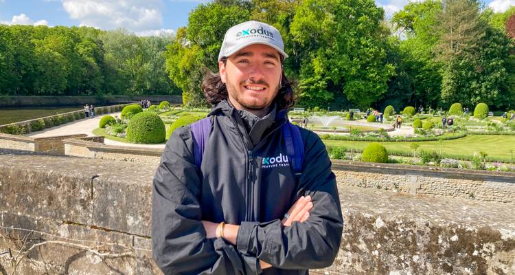 Un guide souriant vêtu d'une veste de marque se tient sur un pont en pierre avec des jardins ornementés et des haies taillées en arrière-plan.