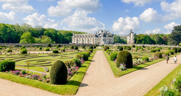 Des parterres de fleurs formels et des allées de gravier guident le regard vers un élégant château de la Renaissance sous un ciel bleu.