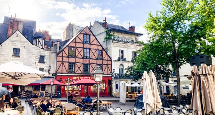Scène animée d'un café en plein air dans une place médiévale à colombages bordée de parasols et de bâtiments historiques.