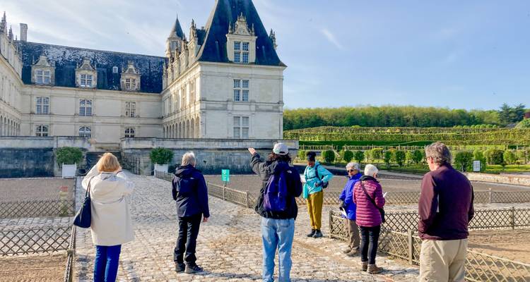 Un guide pointe vers une aile de château ornée tandis que les visiteurs écoutent dans une cour pavée.