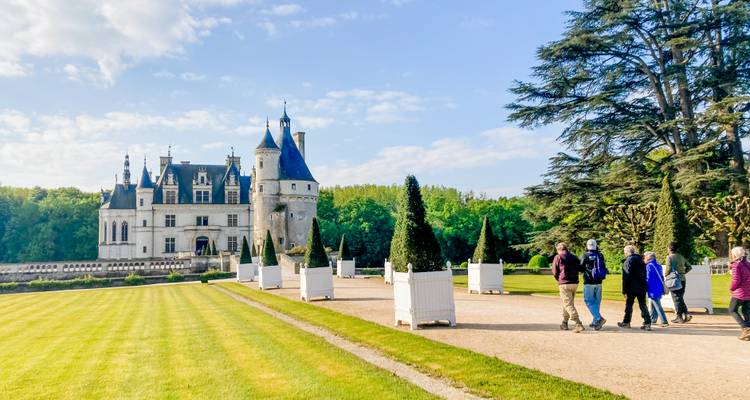 Les voyageurs marchent sur un chemin de gravier vers un château à tourelles encadré par de grands sapins et des pelouses bien entretenues.