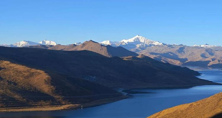 Tranquilo lago alpino que se curva entre colinas áridas y marrones con una cadena de picos nevados bajo un cielo azul despejado.