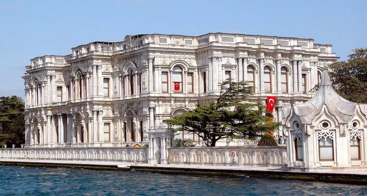 A grand historic palace by the river with Turkish flags and trees.