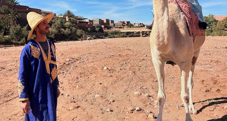 Hombre local con traje tradicional parado al lado de un camello blanco en el lecho seco de un río