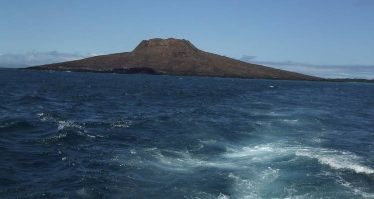 Vue d'une île rocheuse au loin avec une mer agitée et une partie d'un bateau visible.