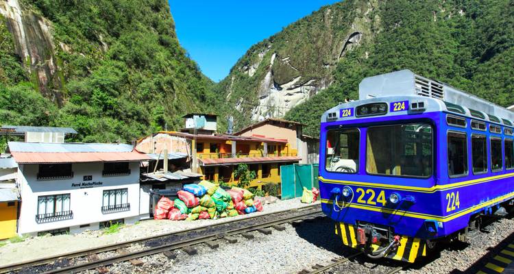 Blauer PeruRail-Zug wartet im Bahnhof Aguas Calientes mit üppiger grüner Bergkulisse und farbenfrohen Ladebündeln in der Nähe