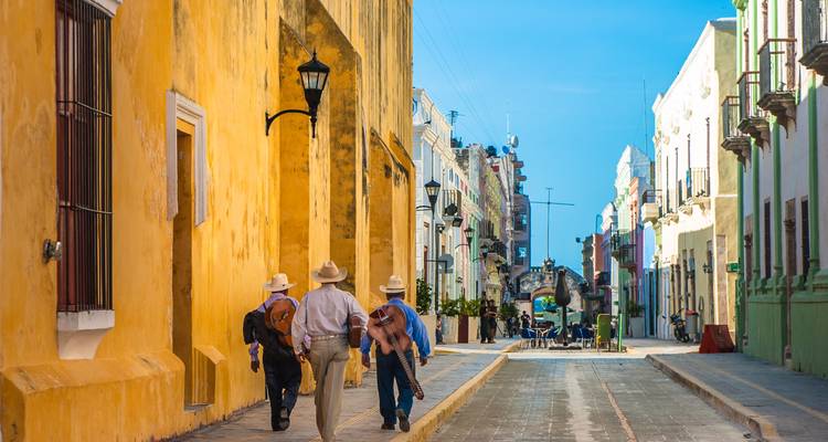 Calle colonial colorida en Campeche con gente local paseando bajo fachadas brillantes usando sombreros de ala ancha.