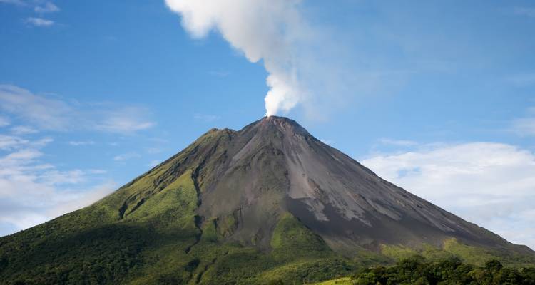 A smoking conical volcano rises above green foothills against a bright blue sky.