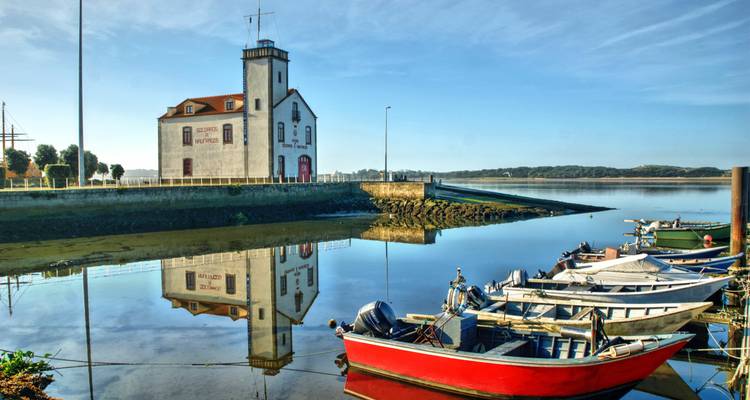 Small fishing boats moored beside a white river-front building reflected in glass-still water under blue sky