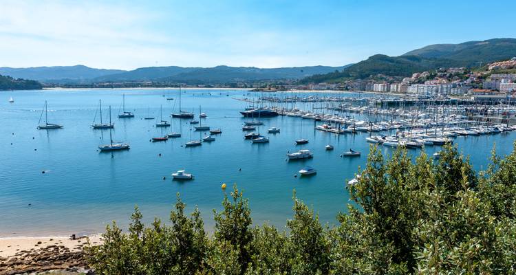 Wide bay filled with anchored sailboats and a marina surrounded by hills and coastal town