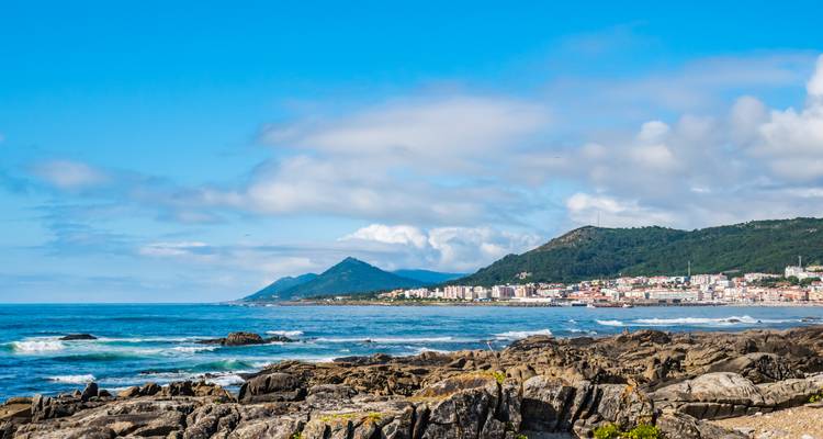 Rocky Atlantic shoreline with coastal town and forested hills beneath bright blue sky and scattered clouds