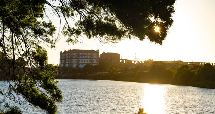 Setting sun glinting through tree branches over a calm river with silhouette of historic buildings and aqueduct